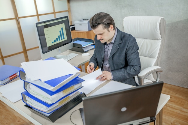 An accountant sitting at a desk reviewing financial documents while consulting a computer screen displaying IRS tax forms. Emphasis on the focused expression on the accountant's face and the organized workspace.